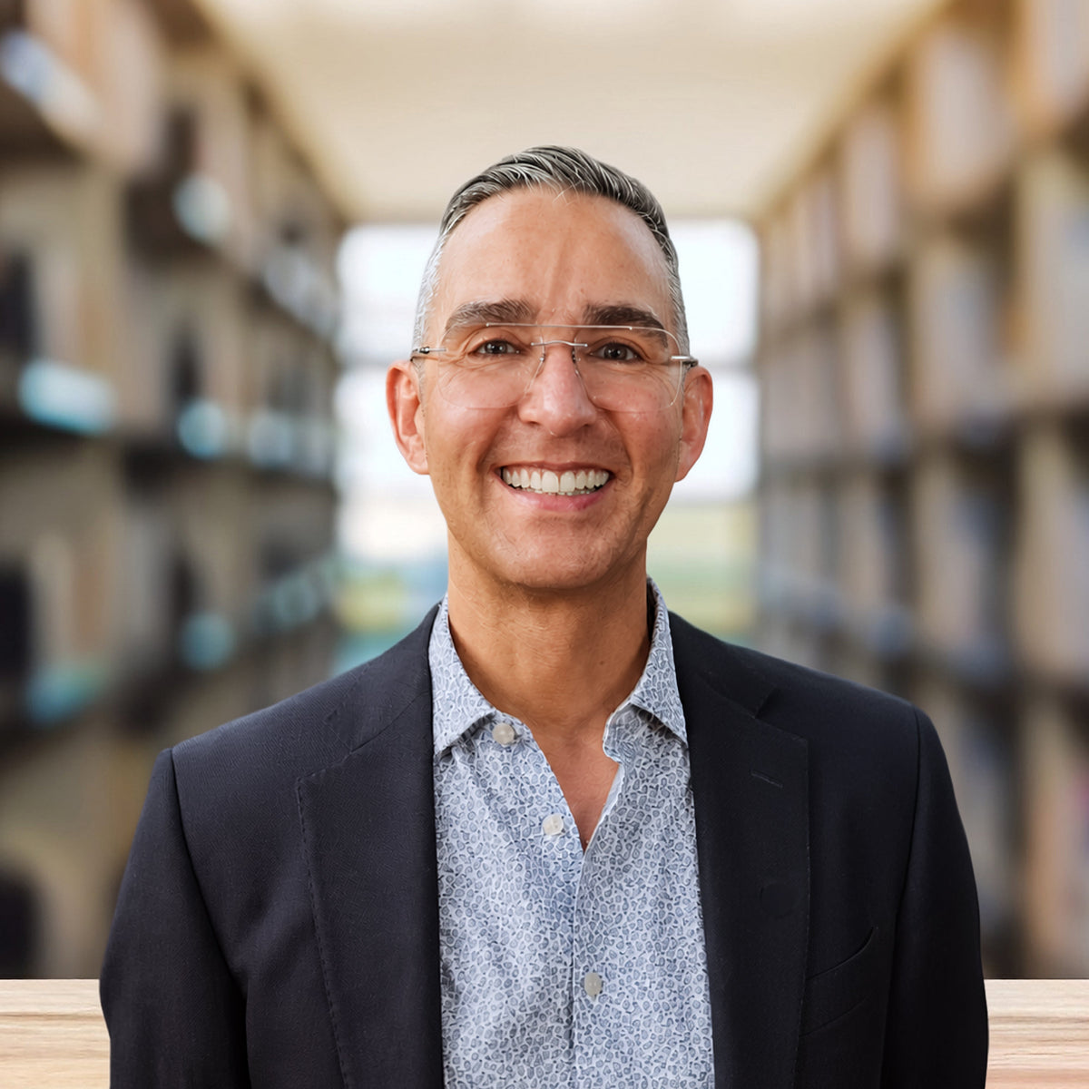 A smiling man with glasses, wearing a dark blazer over a light blue patterned button-down shirt, standing in a library or study with blurred bookshelves in the background.