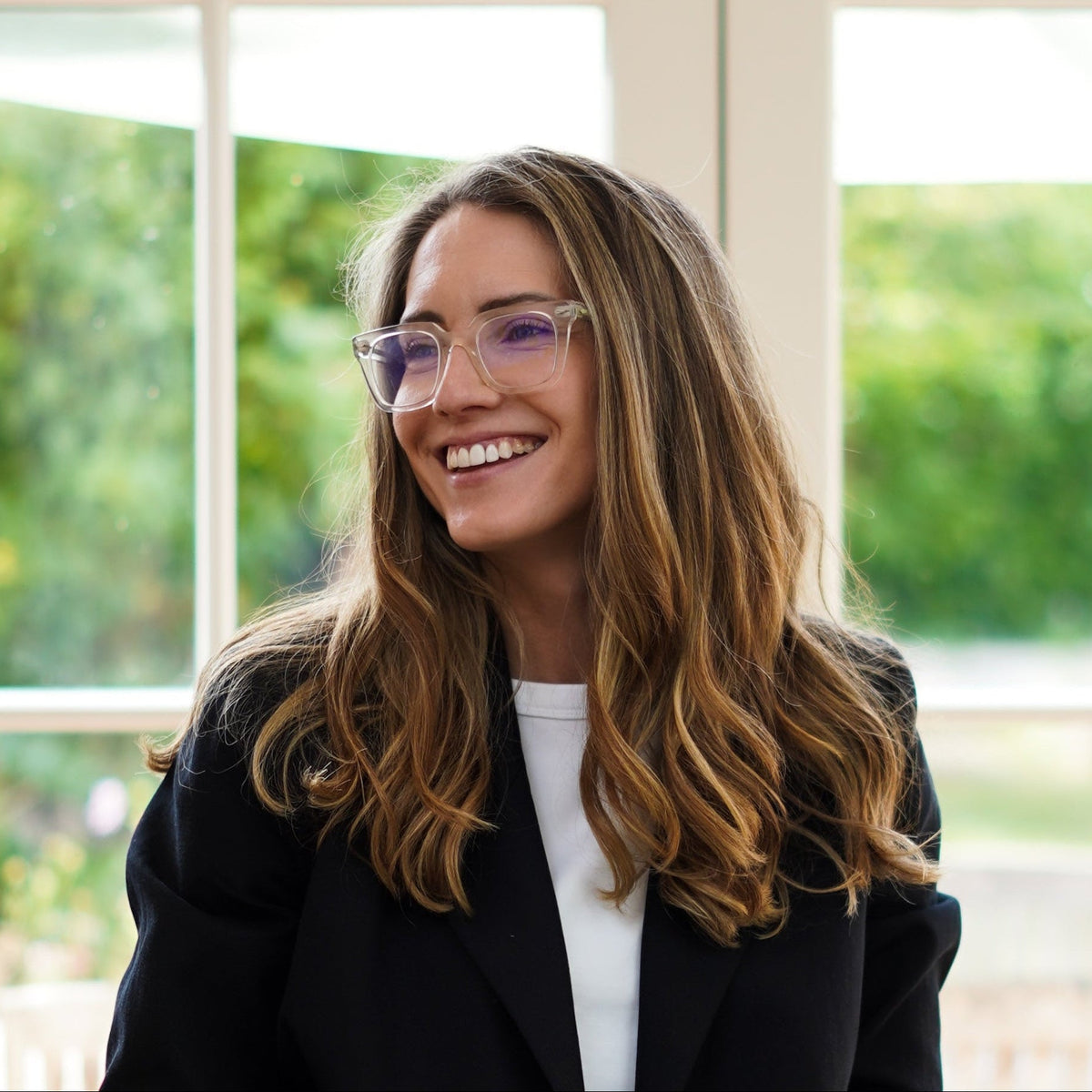 A woman with glasses, wearing a black blazer and blue jeans, sits on a wooden stool holding a laptop in front of large white French doors with a garden view.